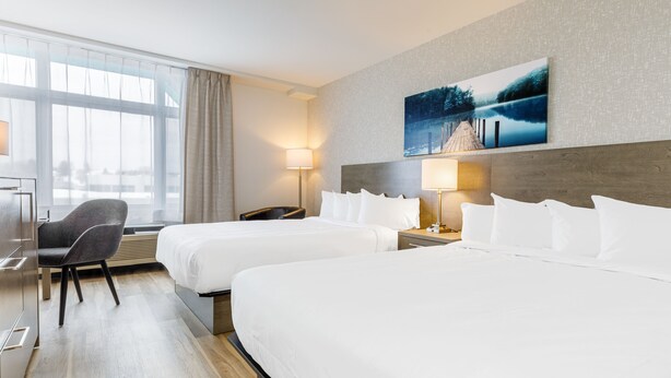A hotel room with two beds, a desk, a chair, and a large window with curtains. - Repotel Henri IV Québec (Québec City)
