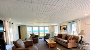 Ein geräumiges Wohnzimmer mit großem Fenster und Blick auf den Strand, ausgestattet mit einer Couch, Sesseln und einem Couchtisch.