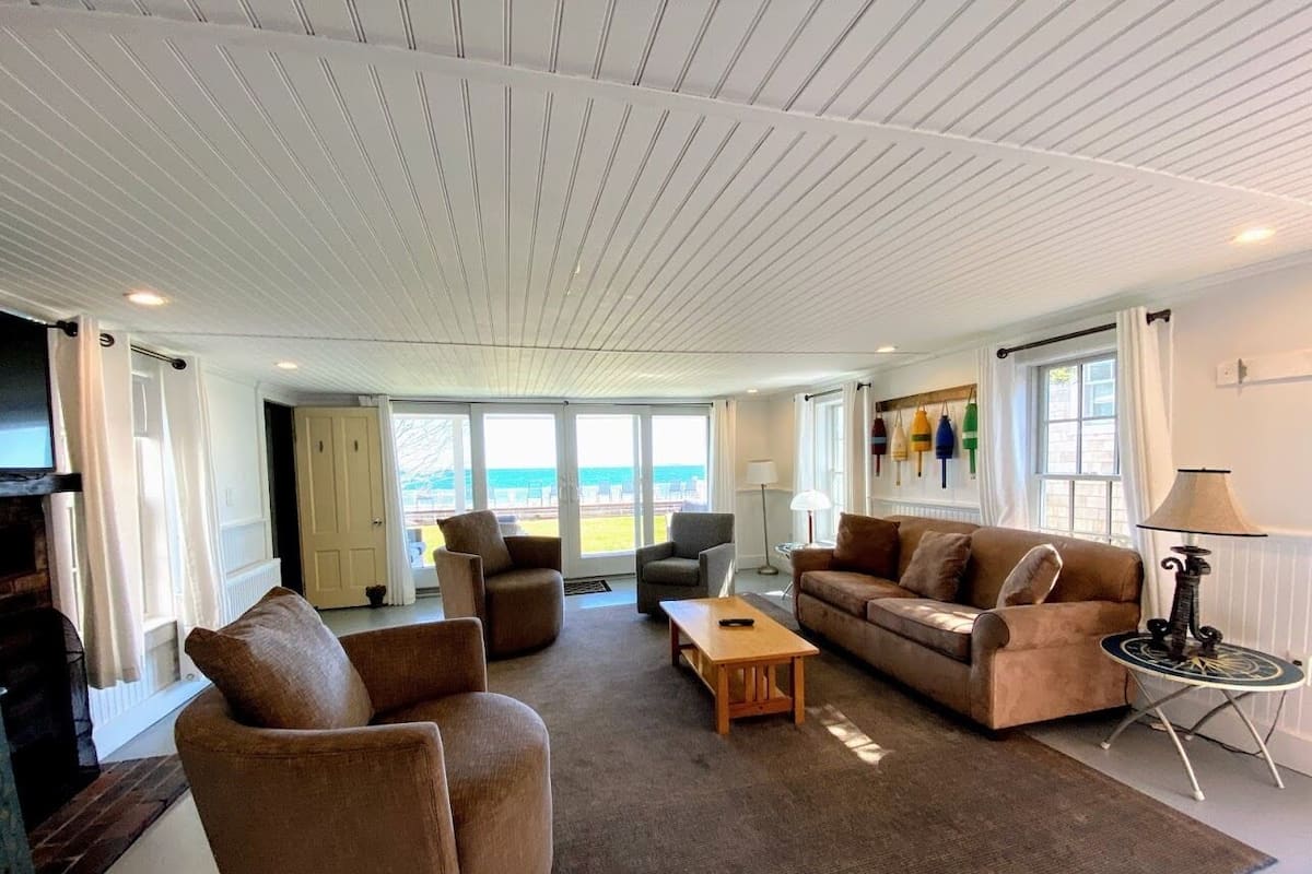 A spacious living room with a large window offering a view of the beach, furnished with a sofa, chairs, and a coffee table.