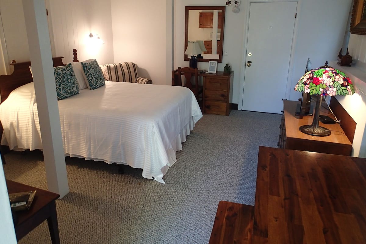 A bedroom with a large bed, a wooden dresser, a desk with a lamp, and a door.