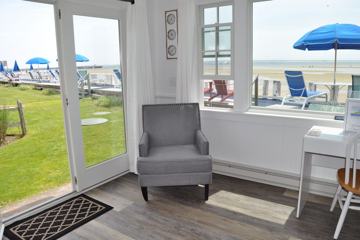 A room with a view of the beach, a grey armchair, a white desk, and a blue umbrella.