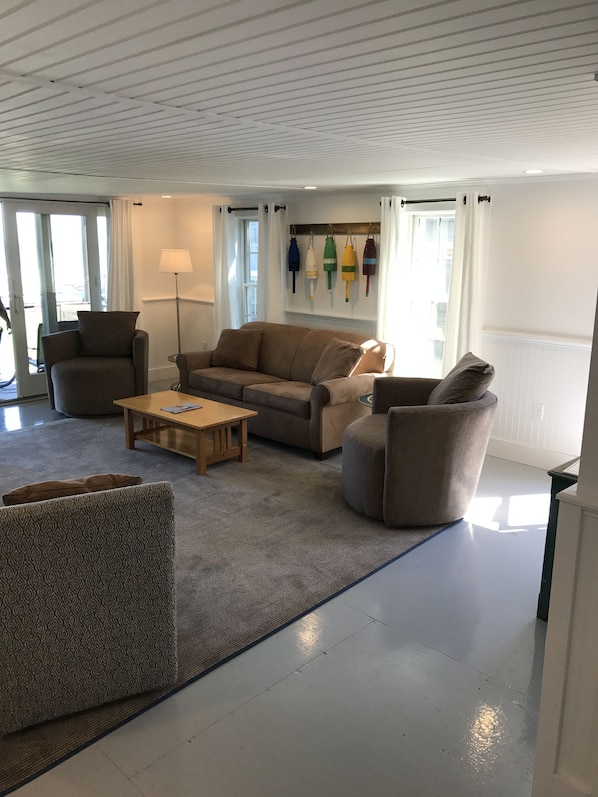 A modern living room with a sofa, armchairs, a coffee table, and a lamp. There is a window with curtains and a wall-mounted shelf with decorative items.