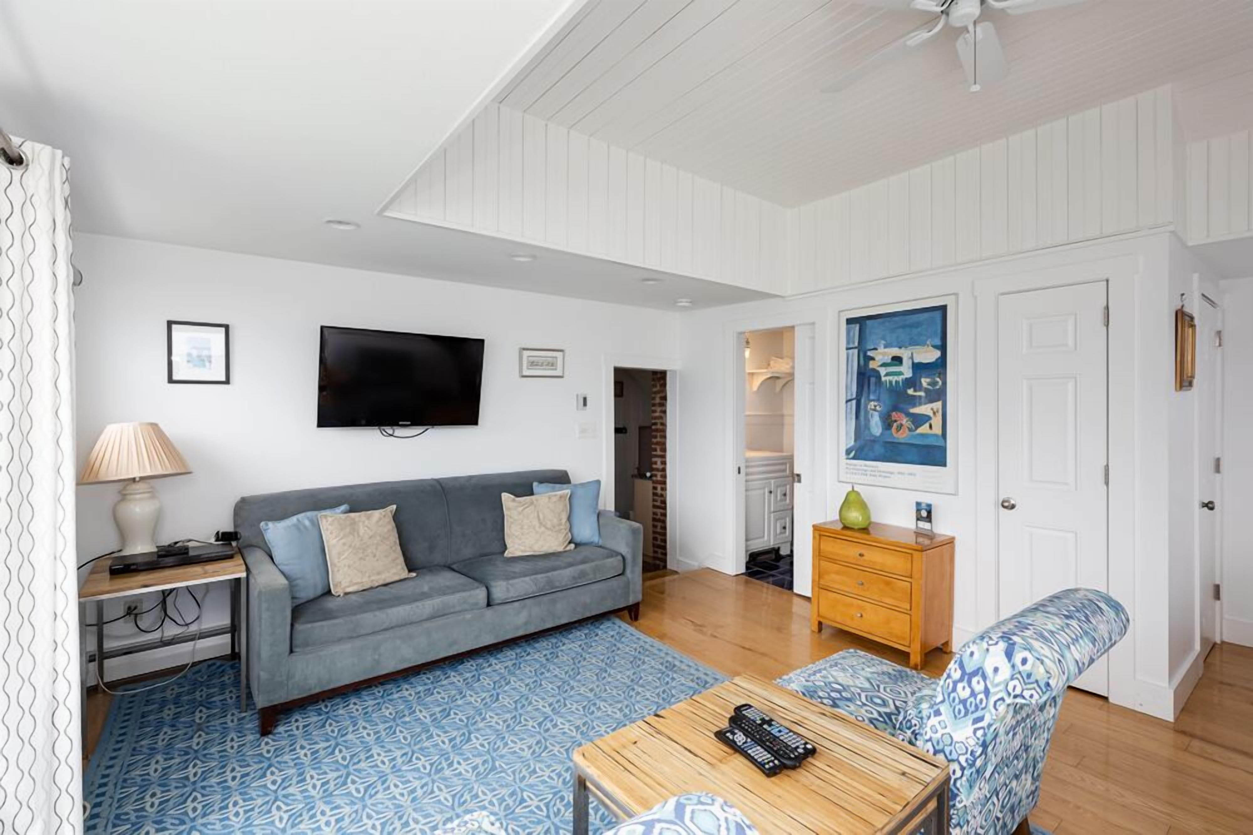 A living room with a grey sofa, a wooden coffee table, a patterned rug, a TV mounted on the wall, and a door leading to a kitchen.