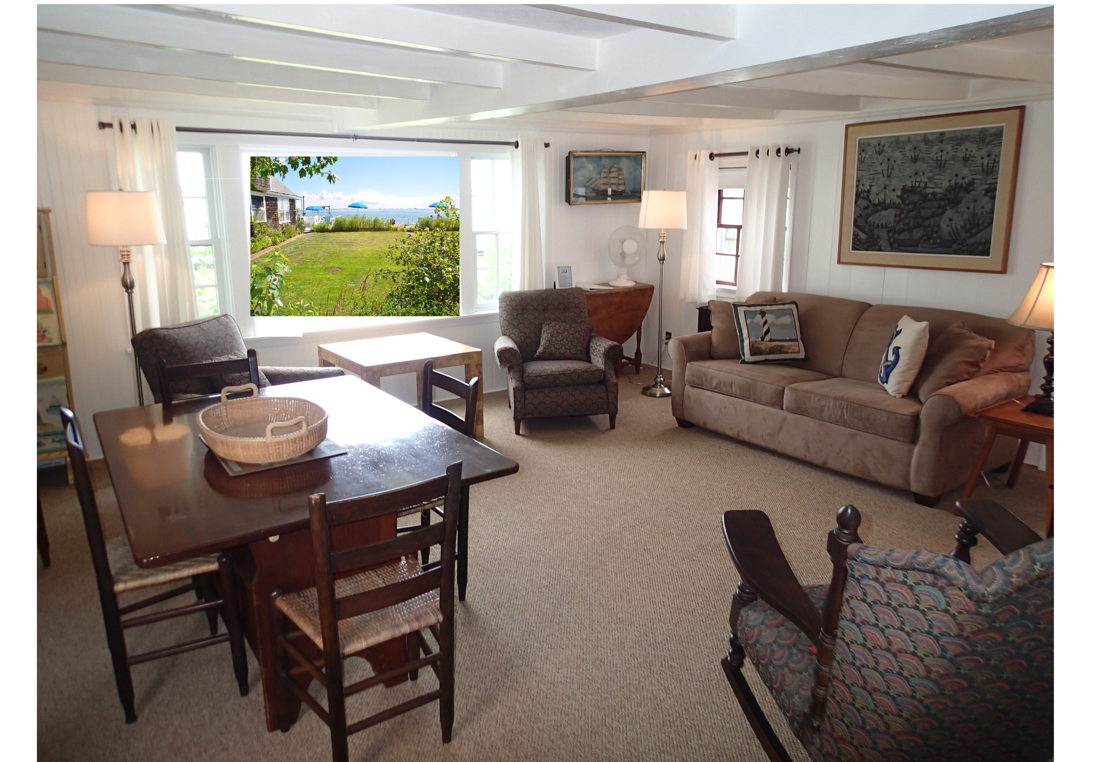 A spacious living room with a large window offering a view of a green landscape, a dining area with a wooden table and chairs, and a comfortable sofa set.