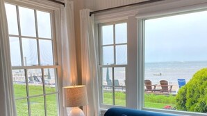 Ein Zimmer mit einem blauen Sofa, einer Lampe und großen Fenstern mit Blick auf einen Strand mit Stühlen und einem Zaun.