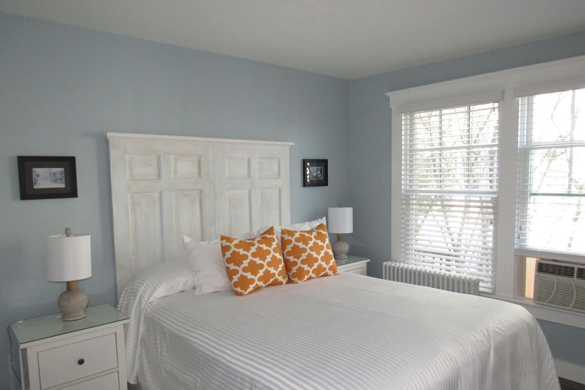 A bedroom with a bed, a nightstand, a lamp, a window with blinds, and a radiator.
