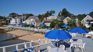 Eine Terrasse direkt am Strand mit weißen Stühlen und blauen Sonnenschirmen, mit Blick auf einen Sandstrand und Wohnhäuser.