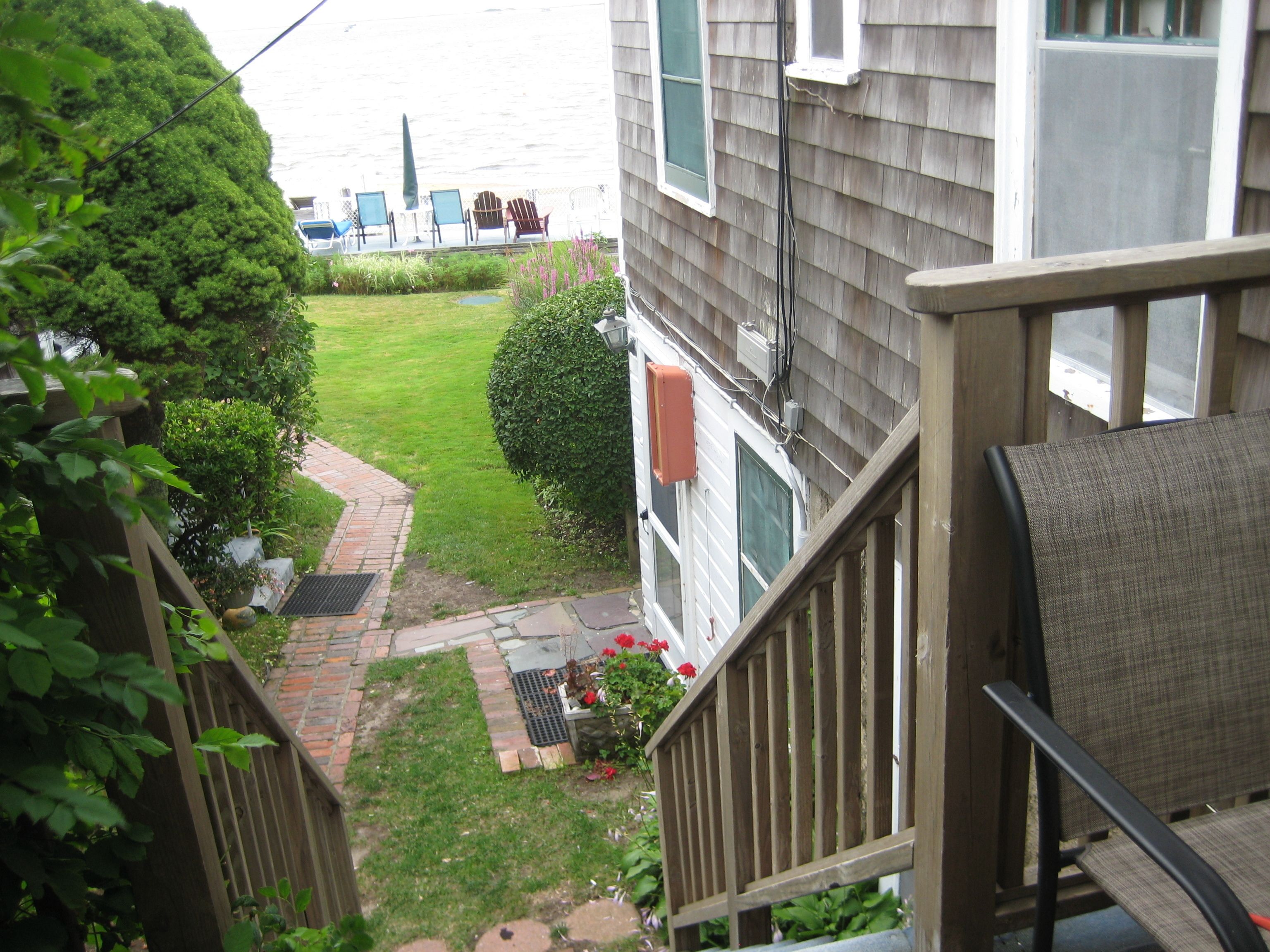A view from a balcony overlooking a small backyard with a lawn, a wooden deck, and a house with a window and a door.