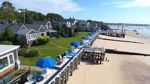 A beachside hotel with a row of sun loungers under blue umbrellas, a wooden pier leading to the beach, and houses in the background. - The Masthead Resort (Provincetown)