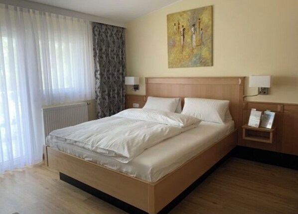 Habitación de hotel con una cama grande, un radiador, una ventana con cortinas, un cuadro en la pared y una mesita de noche con una lámpara.