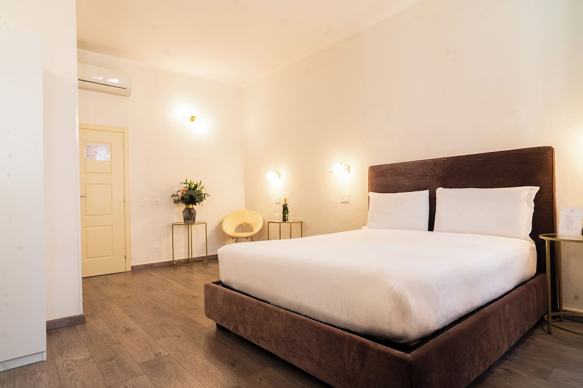 Habitación de hotel con una cama grande, piso de madera, una mesita con un jarrón de flores y una lámpara de pared.