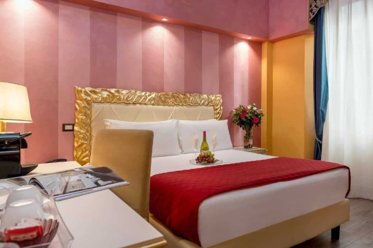 Habitación de hotel con una cama grande, una mesita de noche con lámpara, y una pequeña mesa con una revista y un vaso.