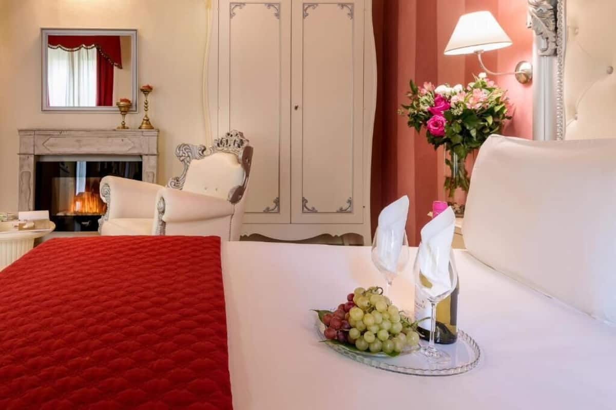 Una habitación de hotel con chimenea, una cama grande con una manta roja, un sillón blanco y un jarrón con flores.
