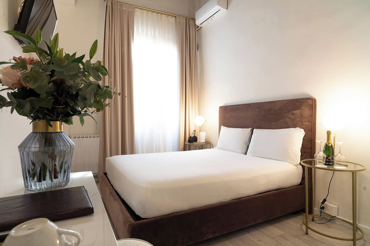 Una habitación de hotel con una cama grande, una mesita de noche con lámpara, un florero con flores y un equipo de aire acondicionado.