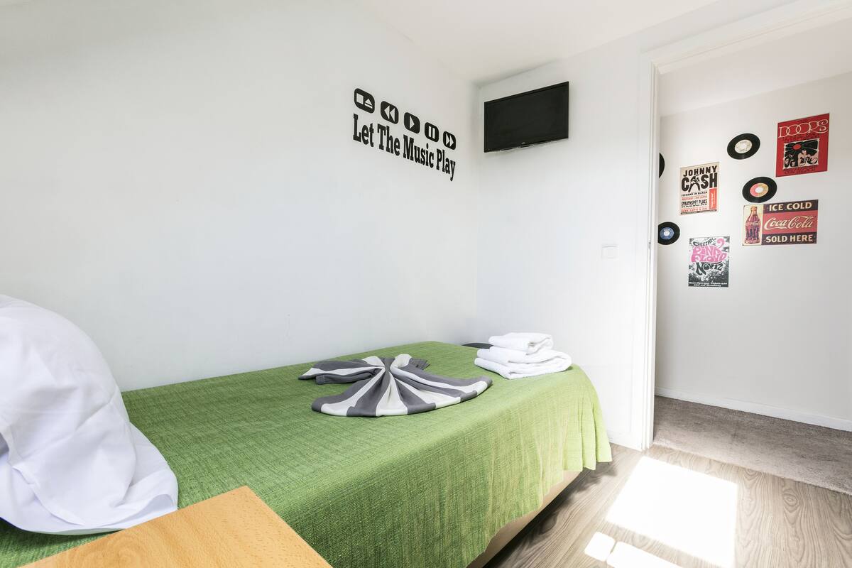 Un dormitorio pequeño y ordenado con una colcha verde, un televisor de pantalla plana montado en la pared y varios discos de vinilo expuestos.