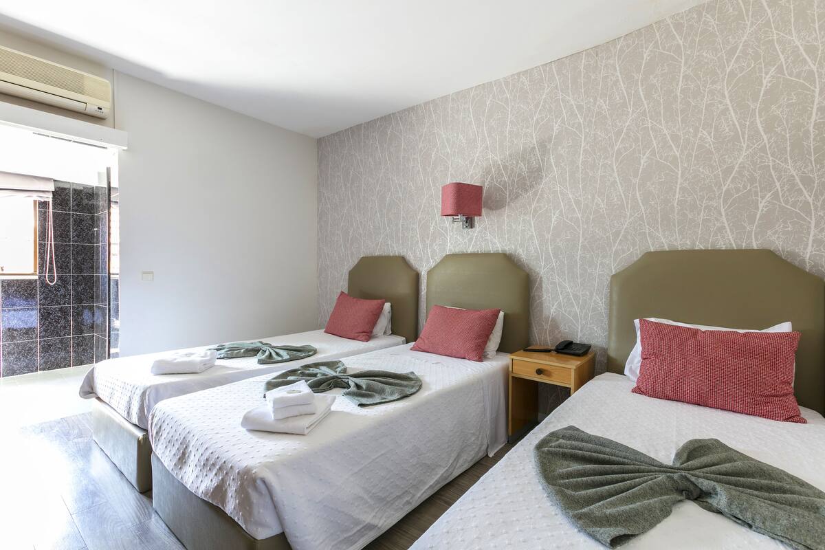 Habitación de hotel con dos camas, una mesita de noche, una lámpara de pared y una ventana con vistas.