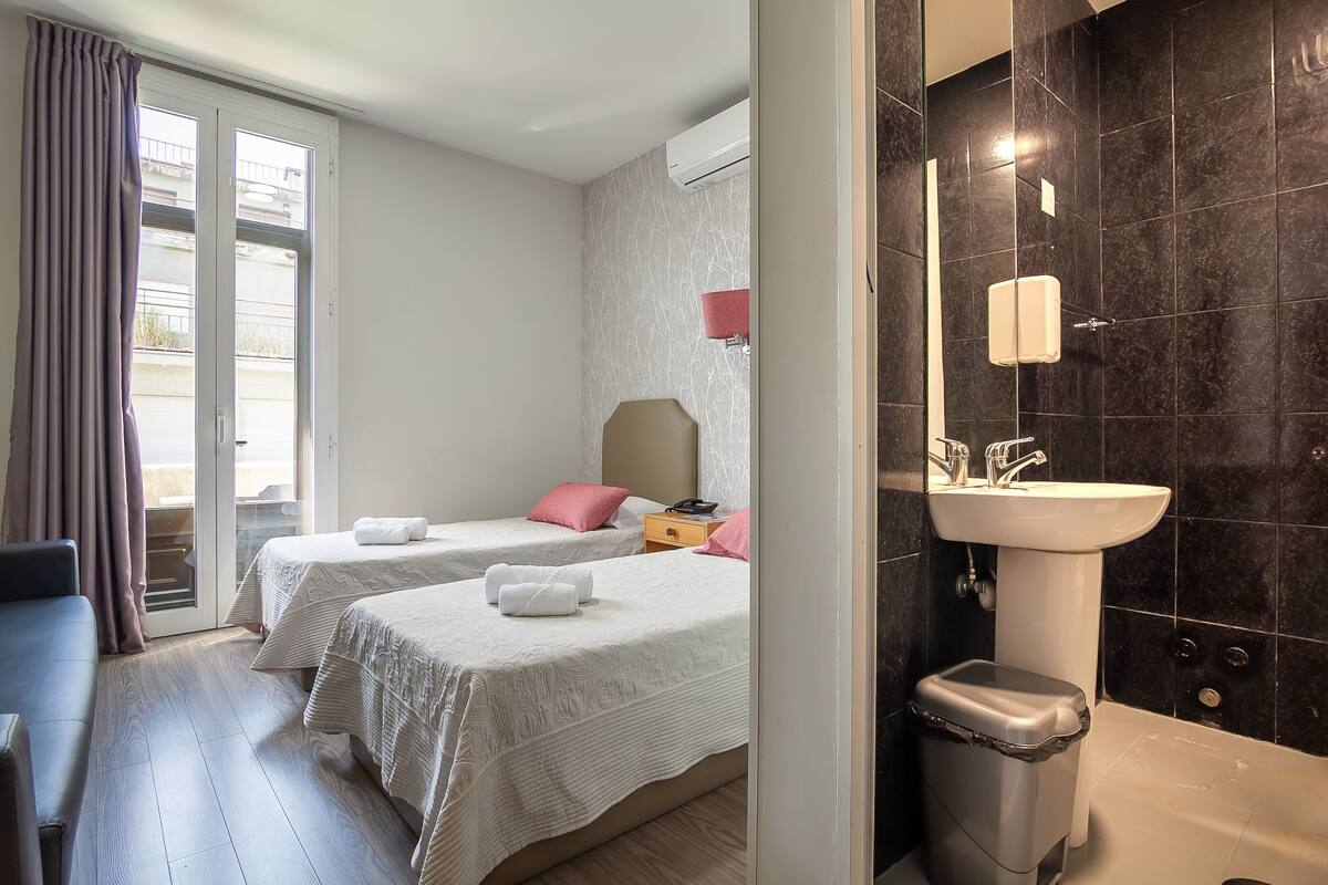 Habitación de hotel con dos camas, un baño con lavamanos e inodoro, y una puerta corrediza de vidrio al exterior.
