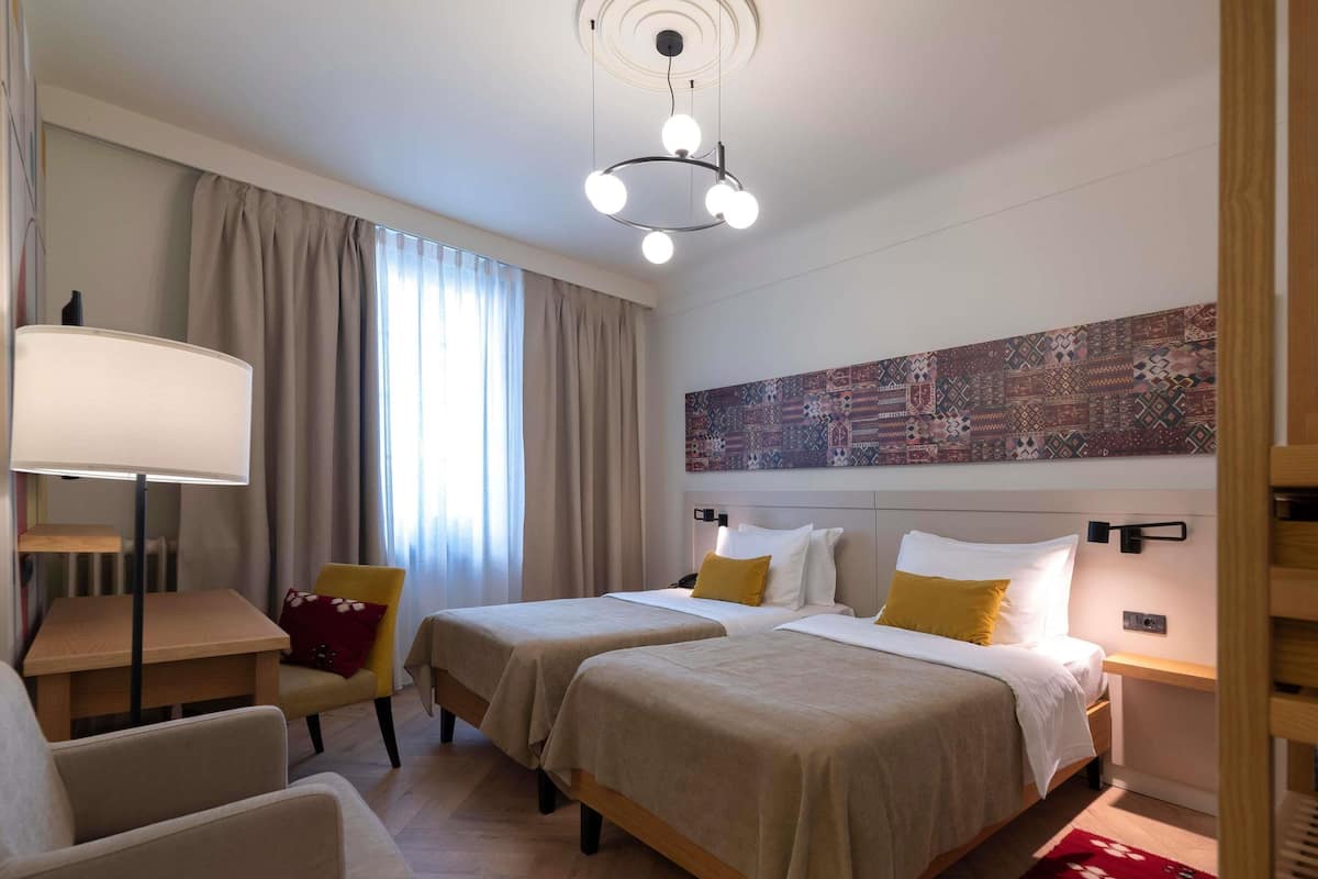 Quarto de hotel com duas camas, uma escrivaninha, uma cadeira, um abajur e uma grande pintura abstrata na parede.
