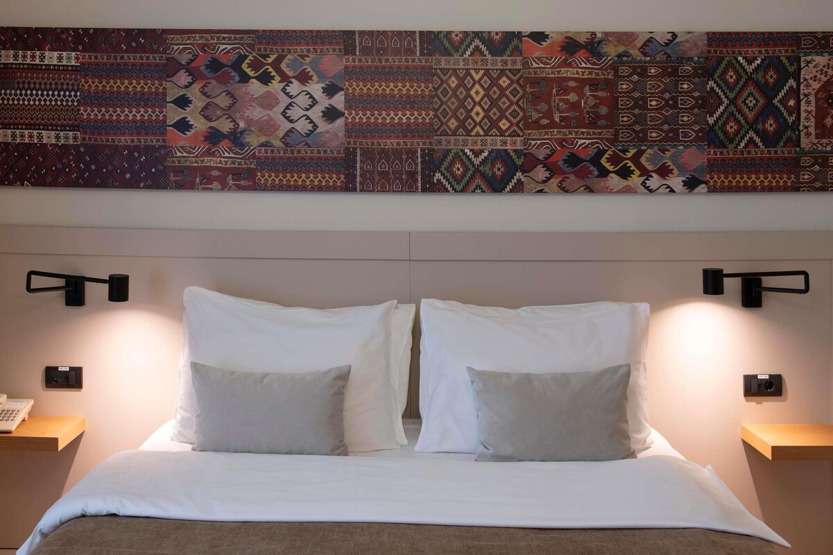 A hotel room with a large bed, two bedside tables, a wall-mounted lamp, and a decorative tapestry.