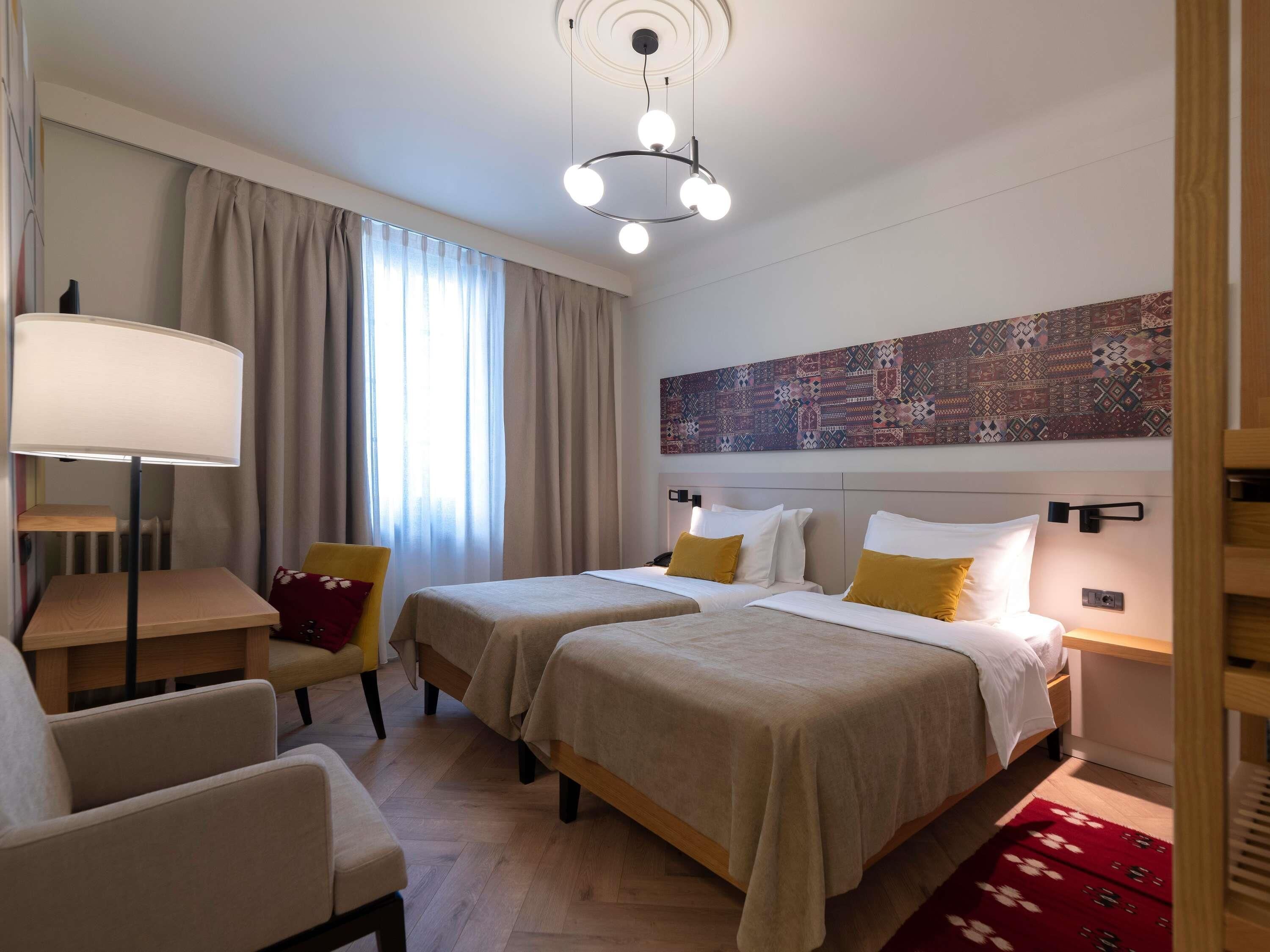 A hotel room with two beds, a desk, a chair, a lamp, and a large abstract painting on the wall.