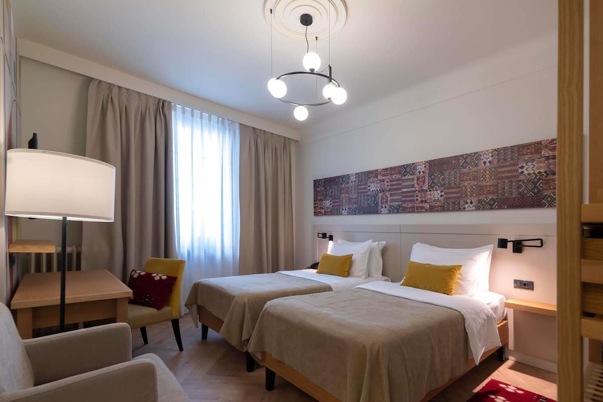 A hotel room with two beds, a desk, a chair, a lamp, and a large abstract painting on the wall.