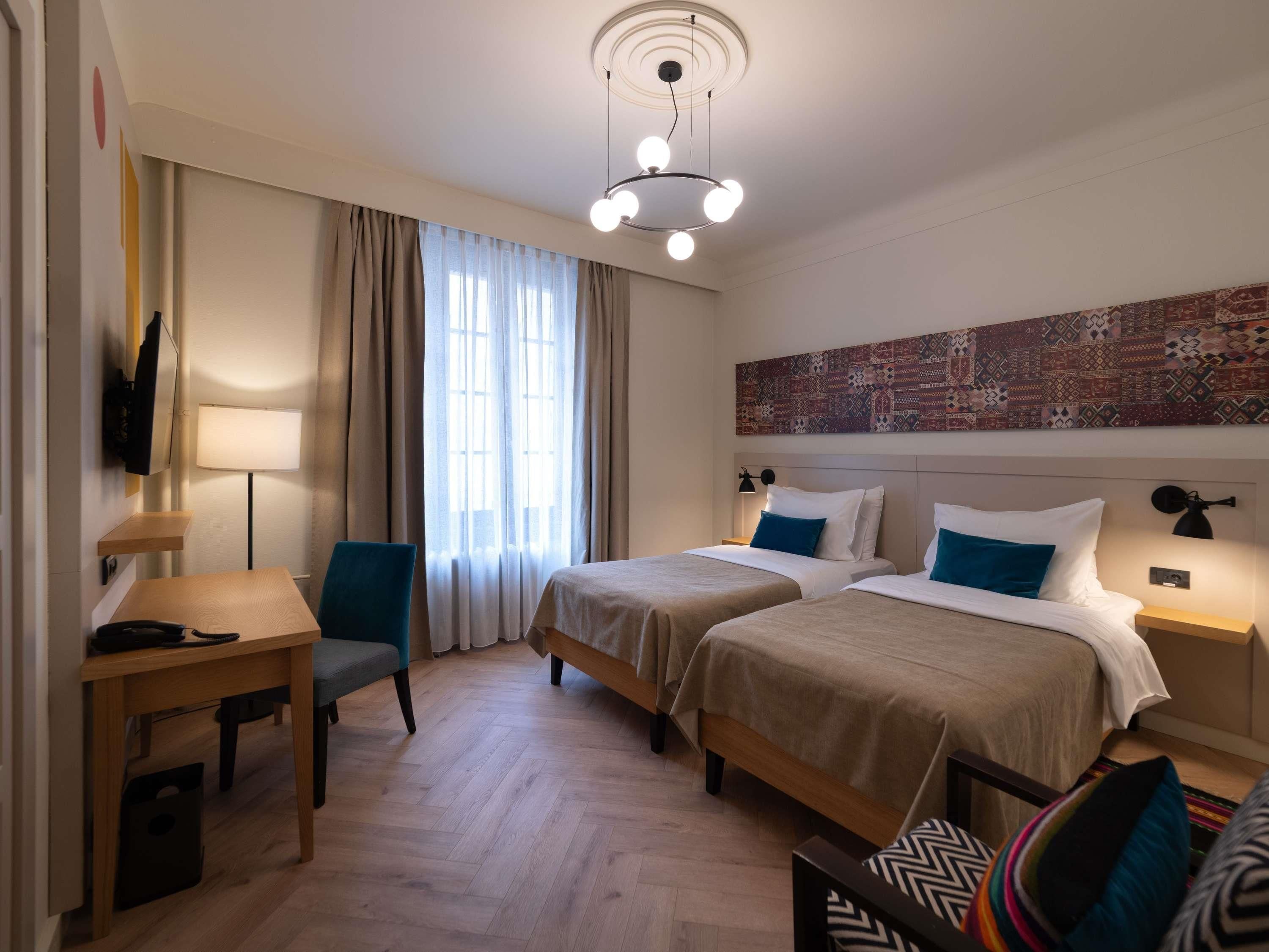 A hotel room with two beds, a desk, a chair, a TV, and a patterned wall.