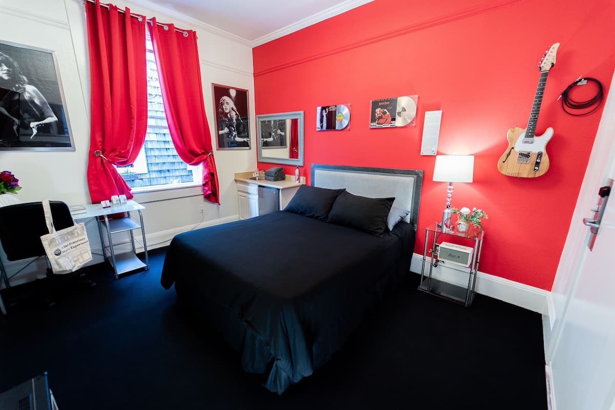Un dormitorio con pared roja, una cama con cubrecama oscuro, una guitarra, un escritorio con lámpara y una ventana con cortinas rojas.