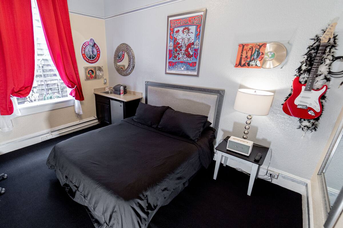 Habitación de hotel con cama, escritorio, silla, ventana con cortinas rojas, lámpara, una guitarra y varios adornos en la pared.