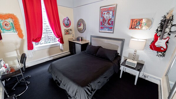 Habitación de hotel con cama, escritorio, silla, ventana con cortinas rojas, lámpara, una guitarra y varios adornos en la pared.