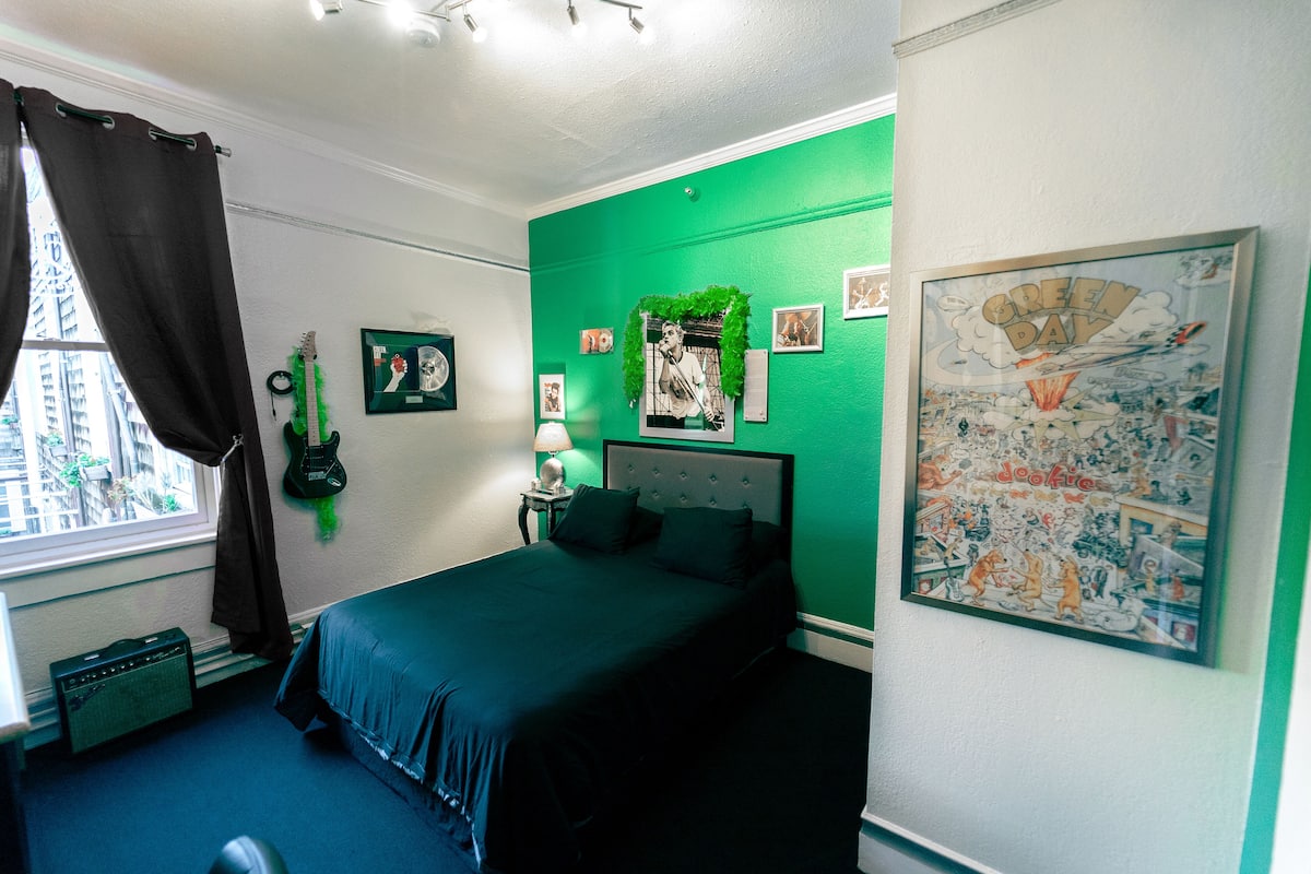 Un dormitorio con una pared verde decorativa, una cama con ropa de cama oscura, una guitarra en la pared y un cartel enmarcado.