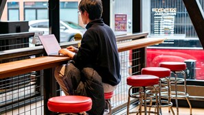 Una persona sentada a una mesa con una computadora portátil, en un café con taburetes rojos y vistas a un vehículo rojo en el exterior.