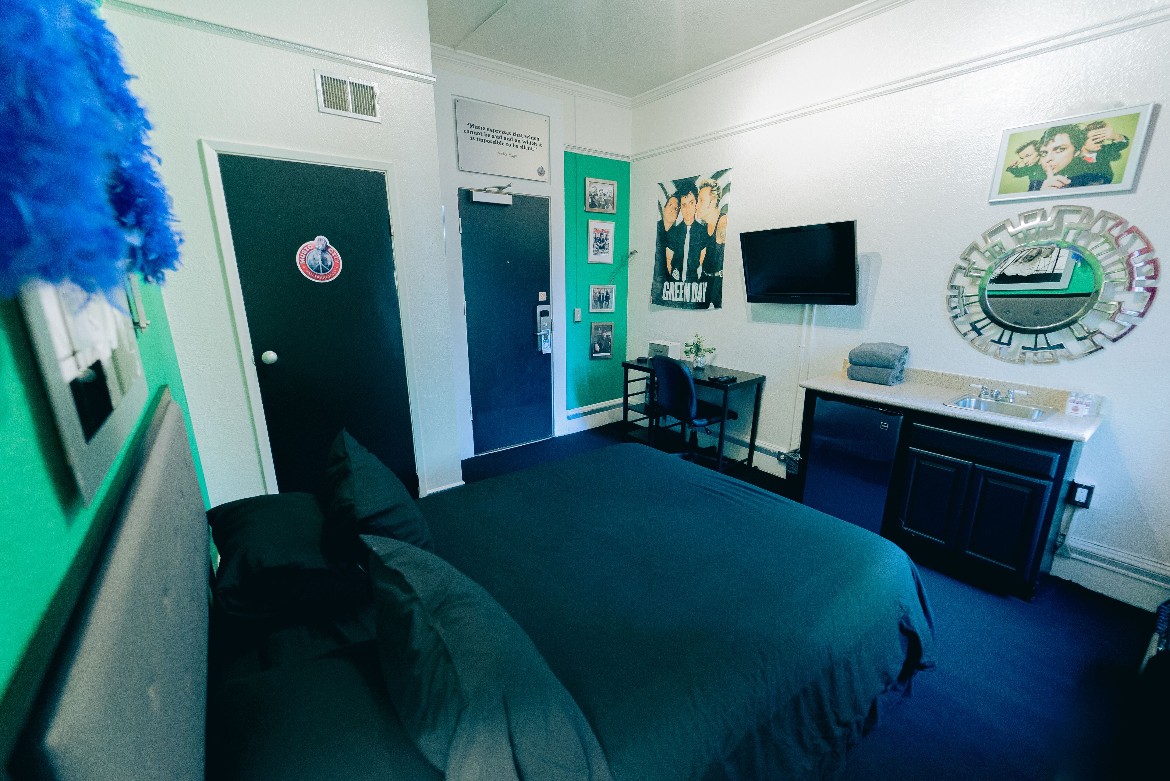 A bedroom with a bed, a desk with a chair, a TV, and a green door.