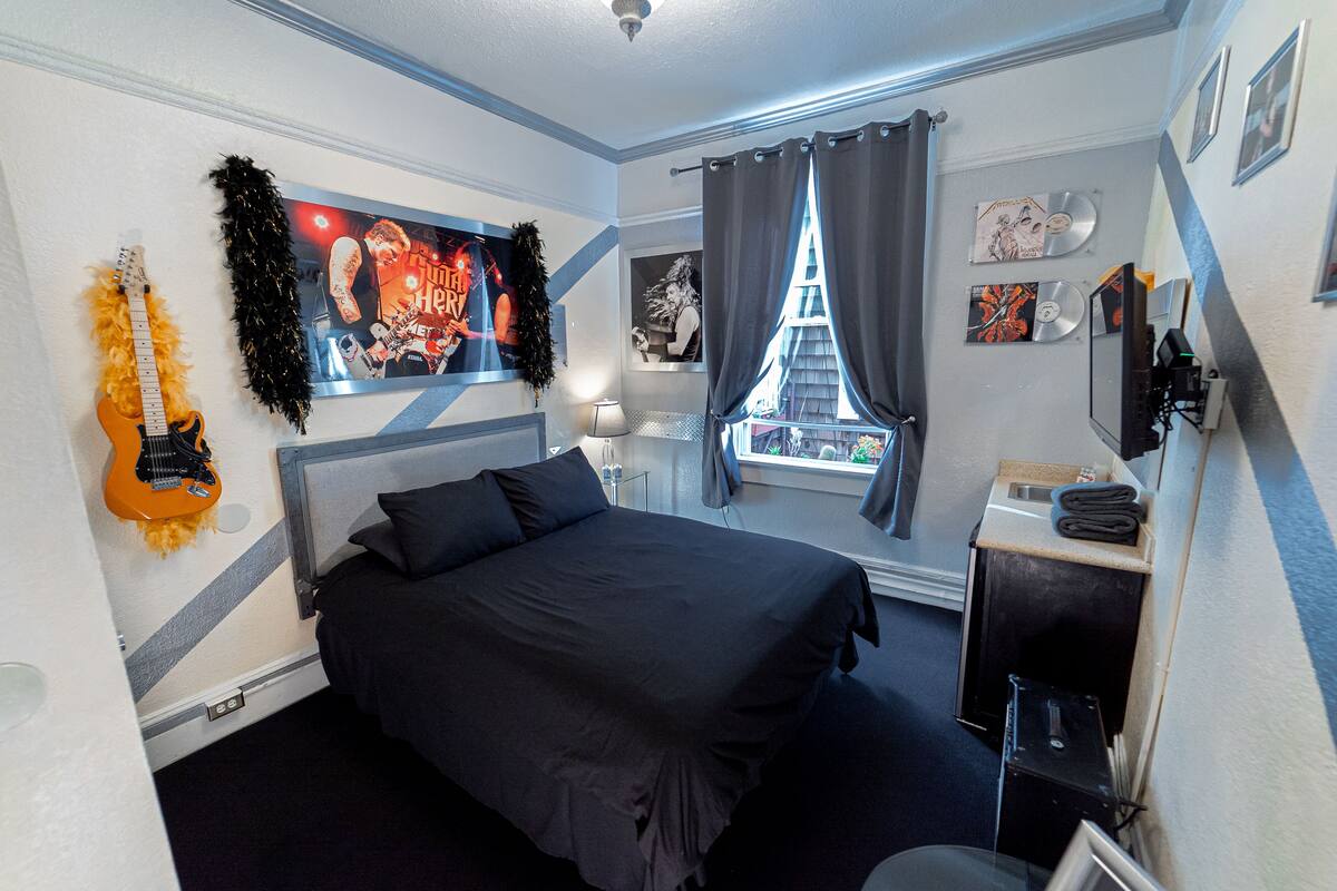 Un dormitorio con cama, una guitarra, una ventana con cortinas y varias decoraciones en la pared, incluyendo carteles y fotografías enmarcadas.