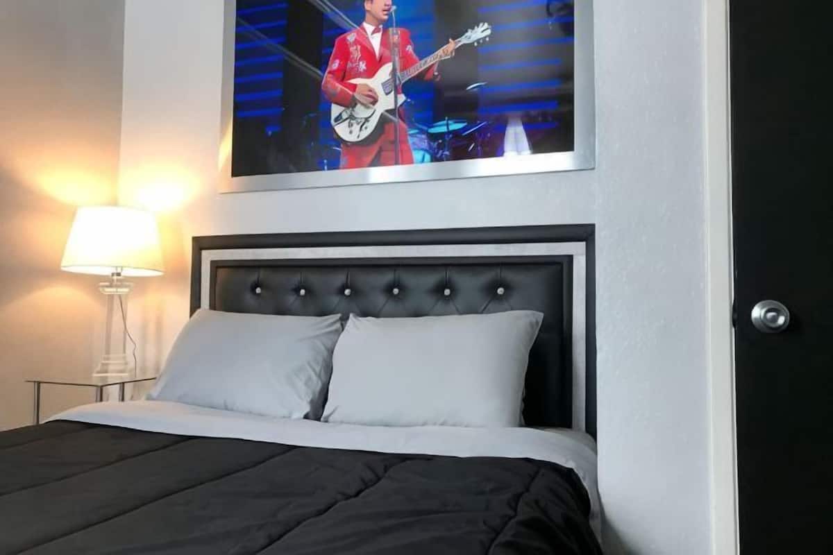 Una cama con cabecera oscura, un almohadón blanco y un cuadro en la pared de una persona tocando la guitarra.