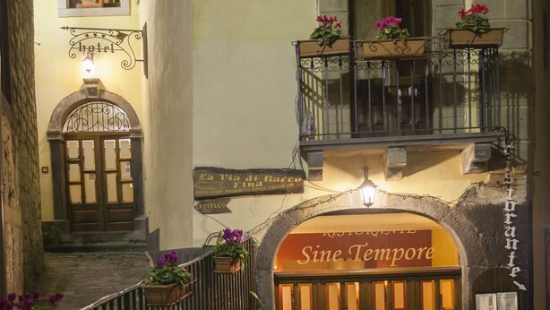 L’entrée d’un hôtel, avec une enseigne portant l’inscription « Sine Tempore », et un balcon agrémenté de plantes en pot.