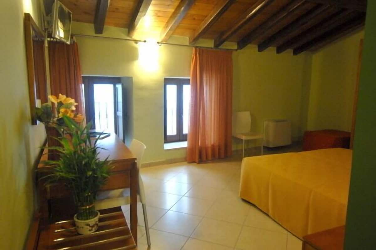 Ein Zimmer mit einem Bett, einem Schreibtisch mit Stuhl, einem kleinen Tisch mit Pflanze und einem Fenster mit Vorhängen.