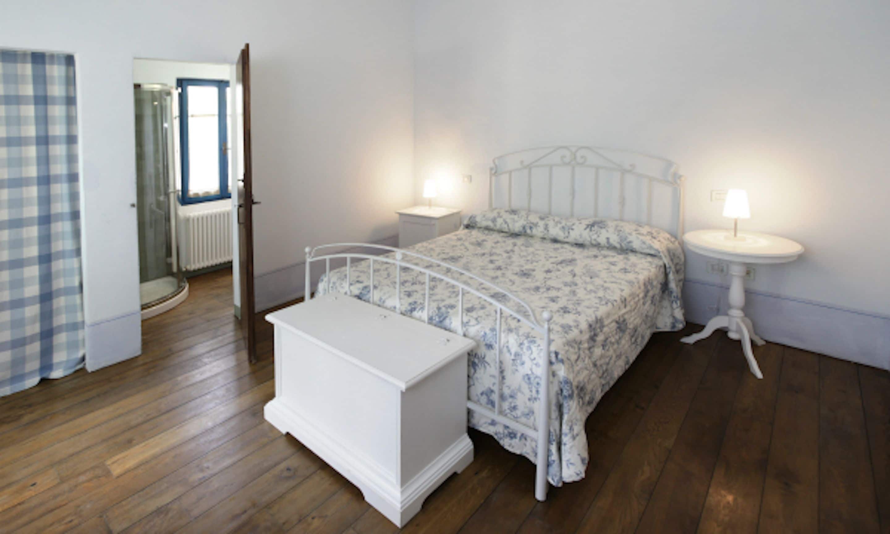 A bedroom with a bed, bedside tables, a nightstand, a small white bench, and a window with a view of a door and a radiator.