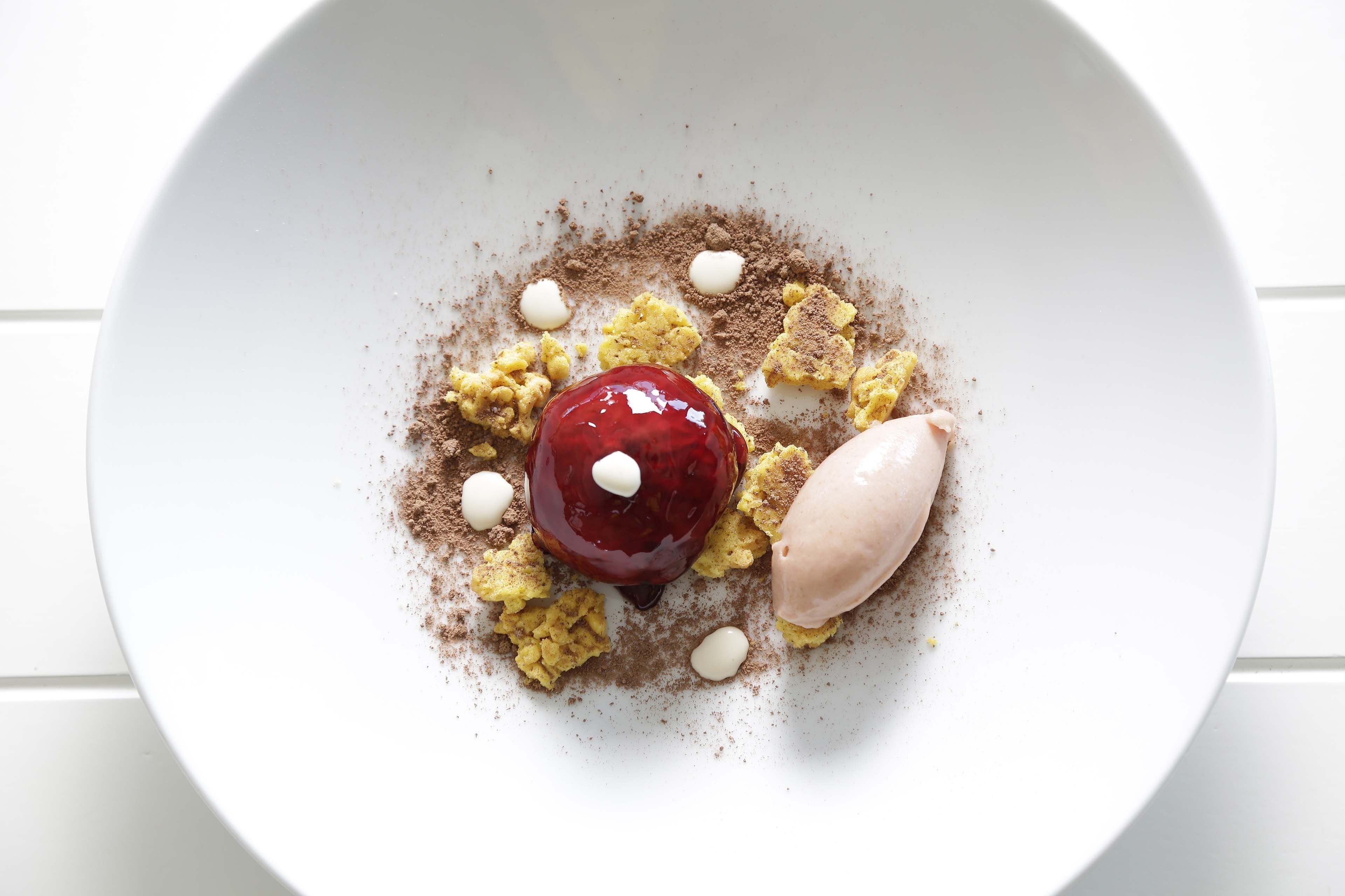 Un postre con una esfera roja, una bola de helado y trozos de pastel desmenuzado sobre un plato blanco.