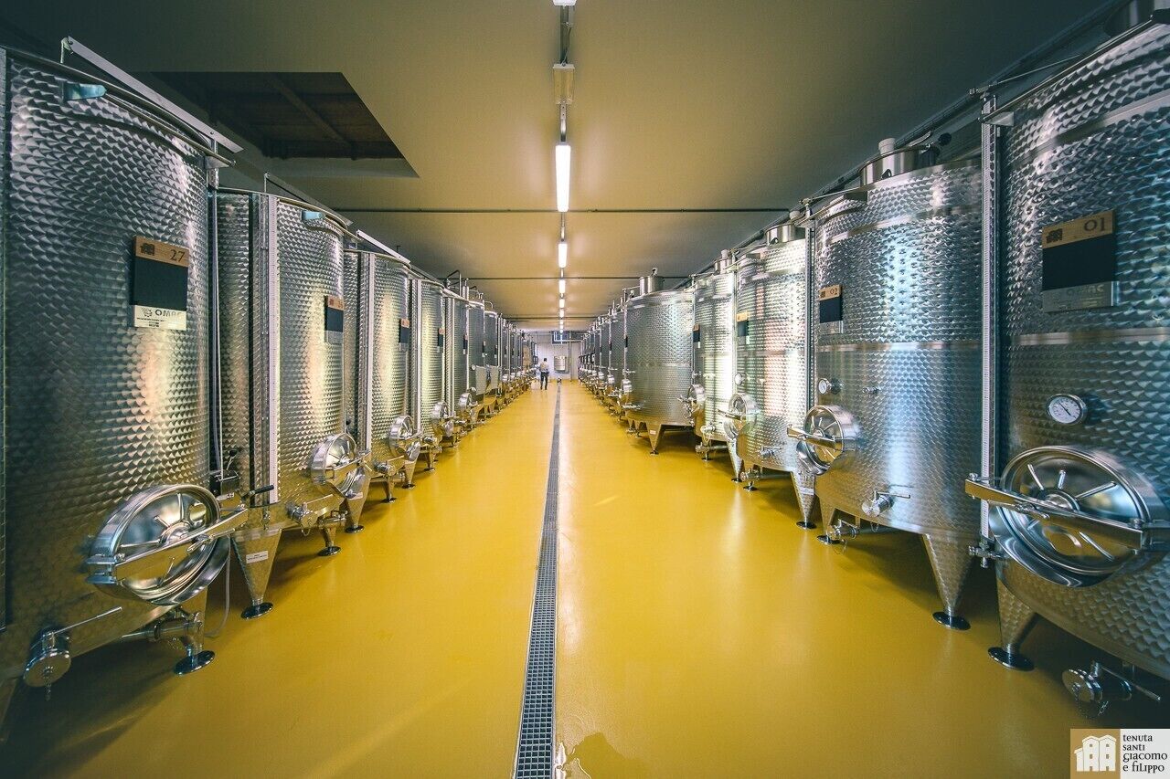 A row of stainless steel fermentation tanks in a facility with a yellow floor and a central walkway.