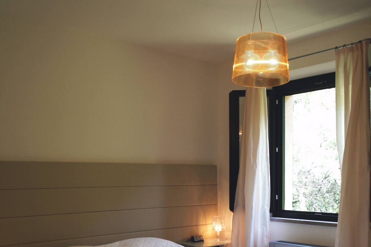 Une chambre à coucher avec un grand lit, une table de chevet, un luminaire suspendu et une fenêtre avec des rideaux.