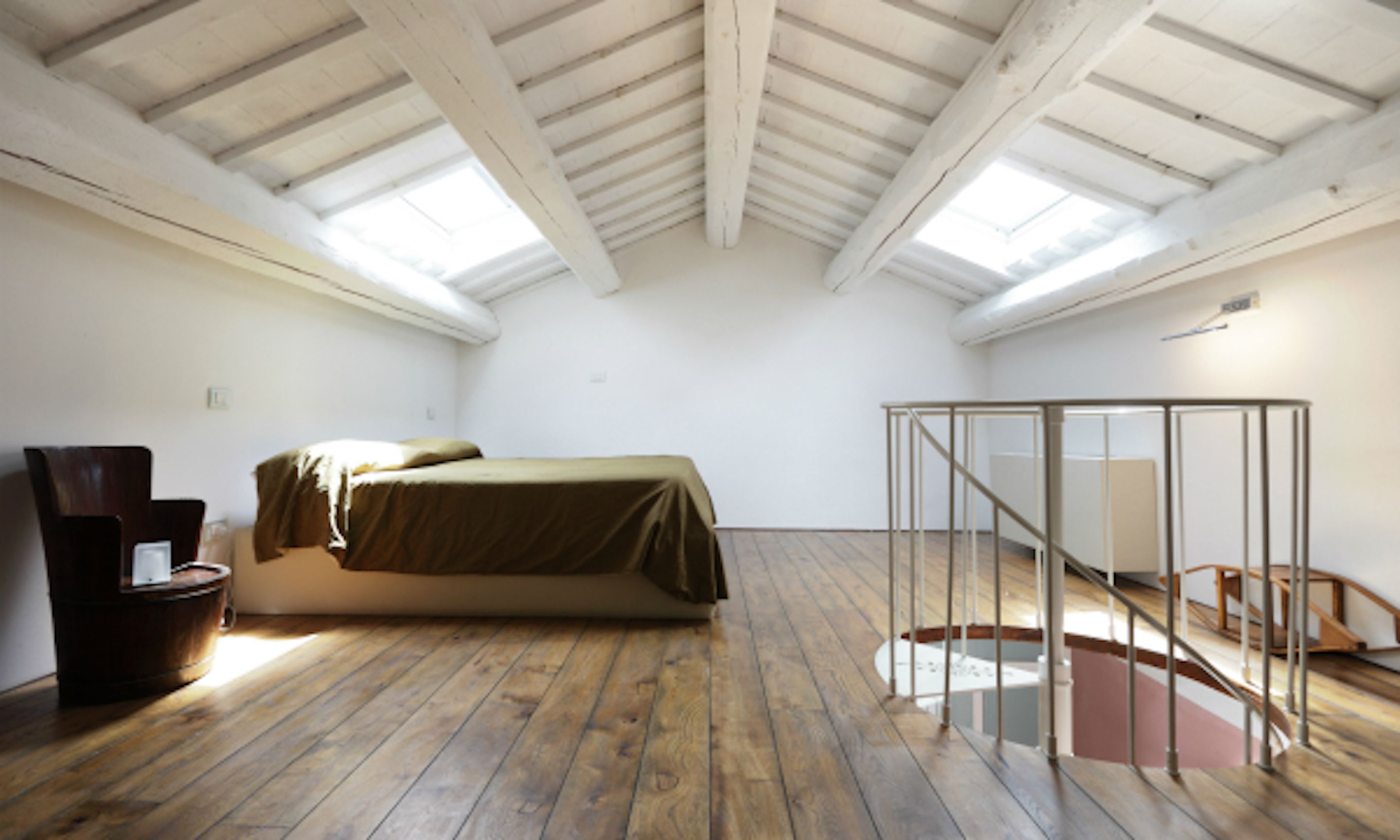 A high-ceilinged room with a wooden floor, a bed, a chair, and a glass partition.