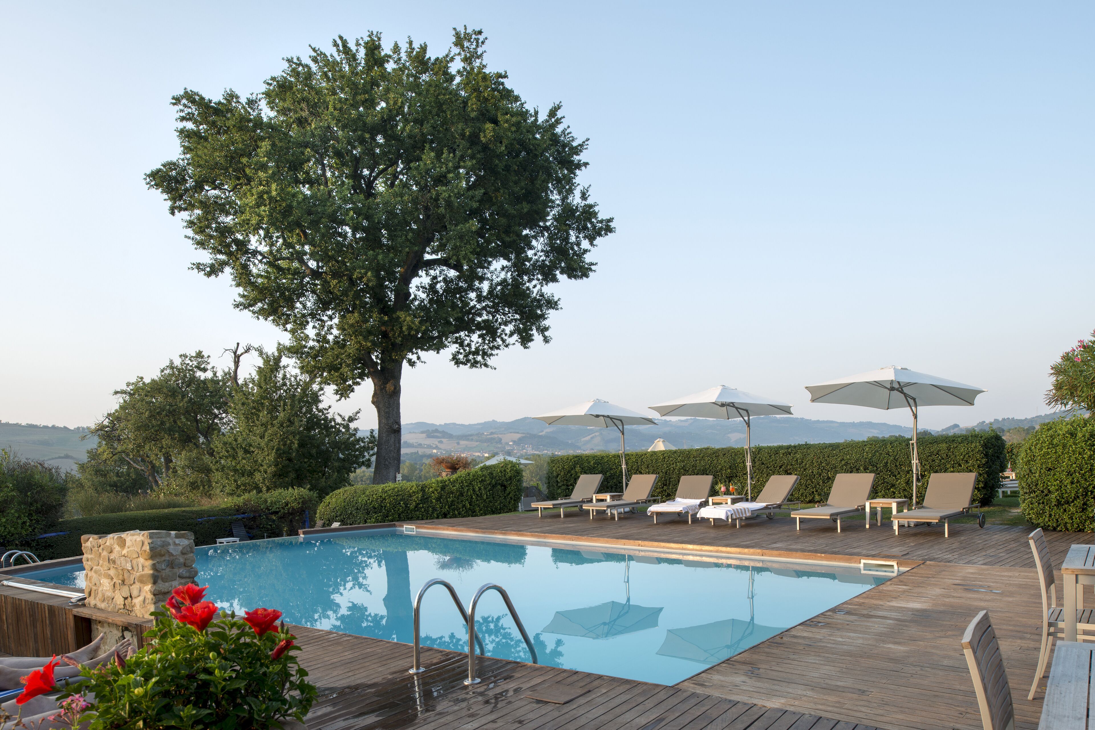A pool area with lounge chairs and umbrellas, surrounded by greenery and a view of distant mountains.