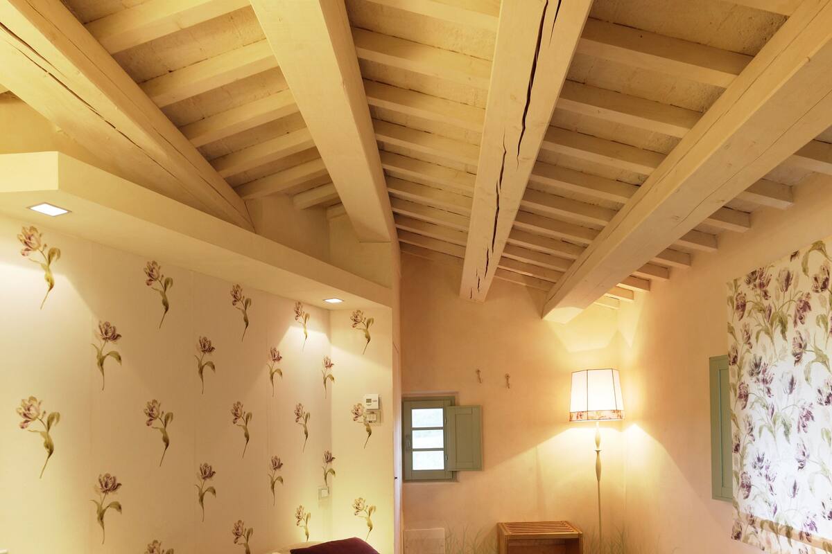 Une chambre avec un plafond en bois, un papier peint à motifs floraux, un lit recouvert d’une courtepointe à fleurs, une table de chevet et une lampe.