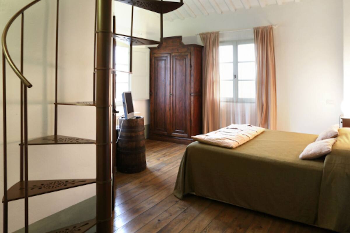Une chambre avec un plancher en bois, un lit recouvert d’une couverture verte, une armoire en bois et un escalier en colimaçon.