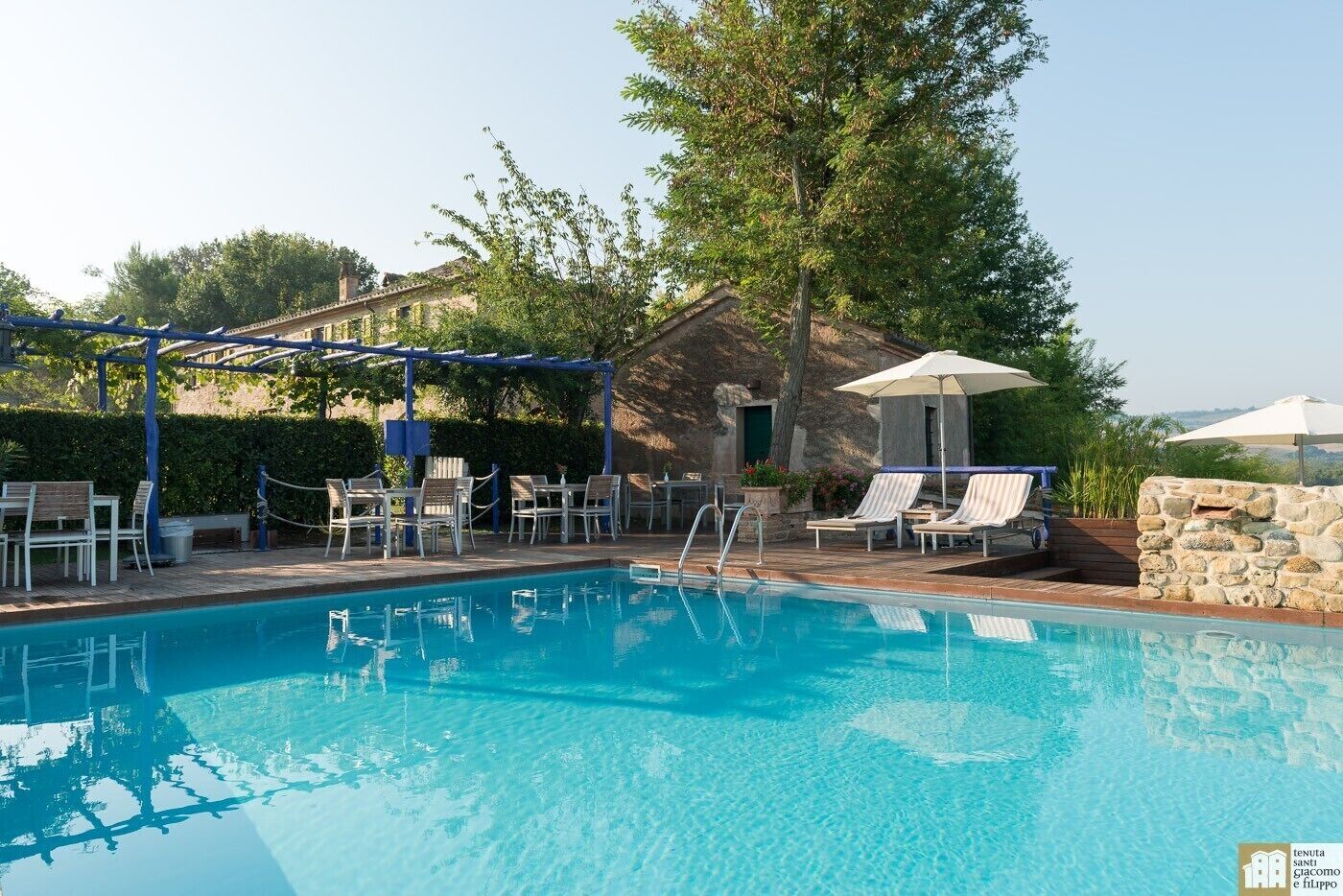 Una piscina con sillones de descanso y sombrillas, rodeada de muros de piedra y vegetación.