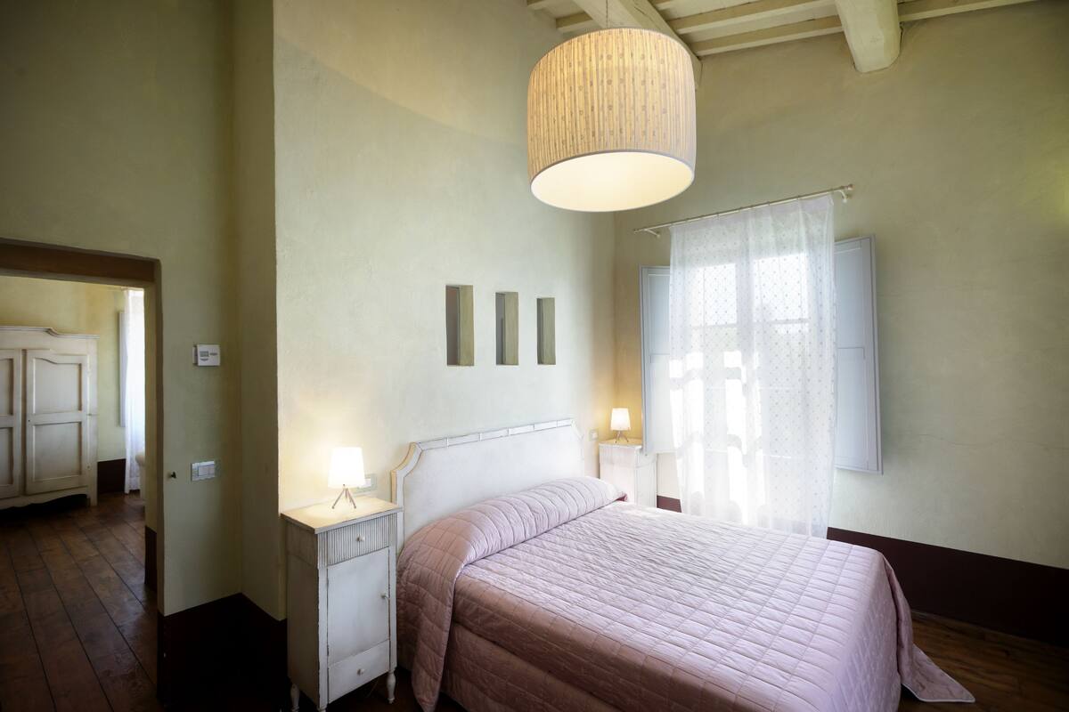 Une chambre à coucher avec un lit, des lampes de chevet, un luminaire suspendu et une fenêtre avec des rideaux.