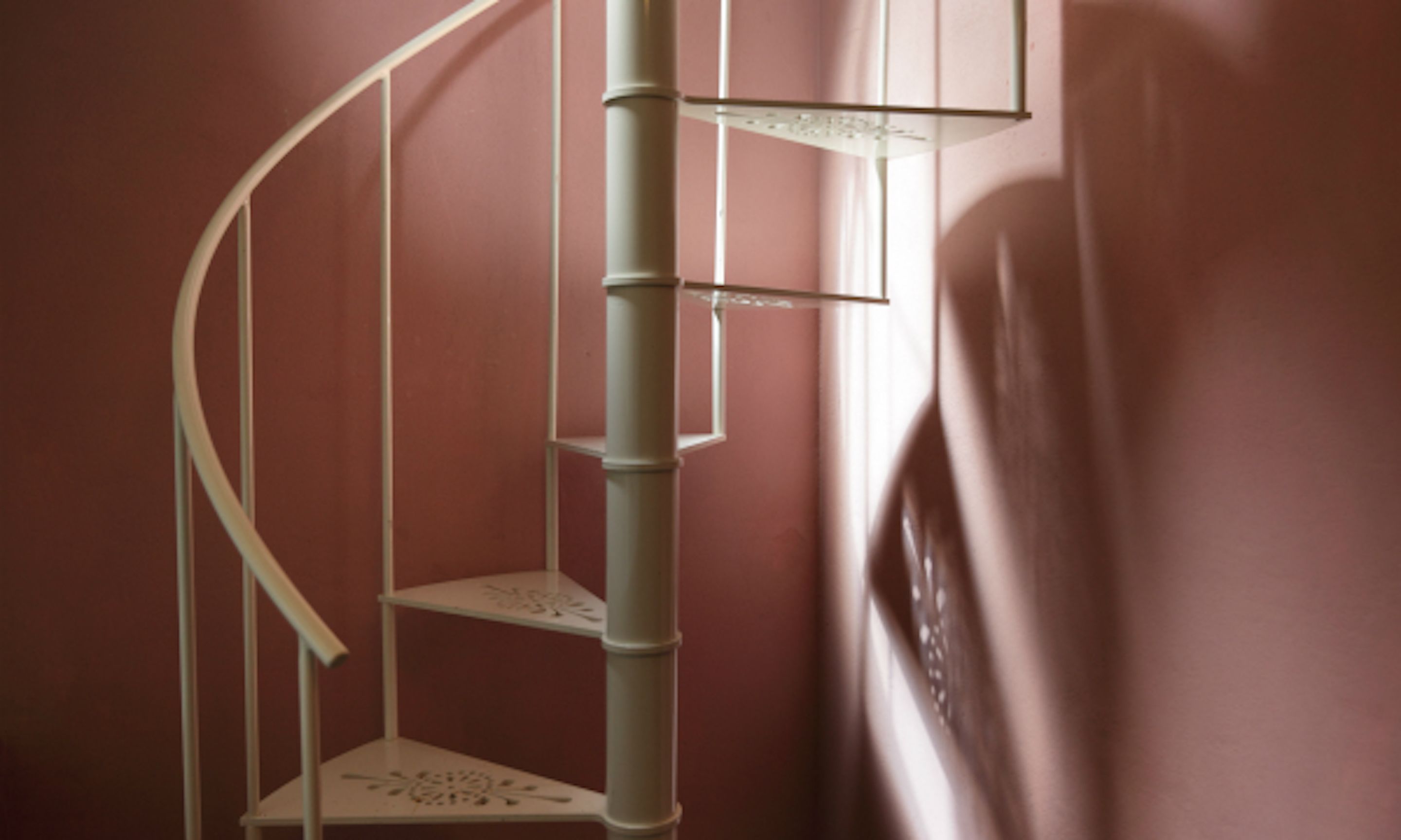 Escada em espiral com corrimão de metal branco em frente a uma parede rosa.
