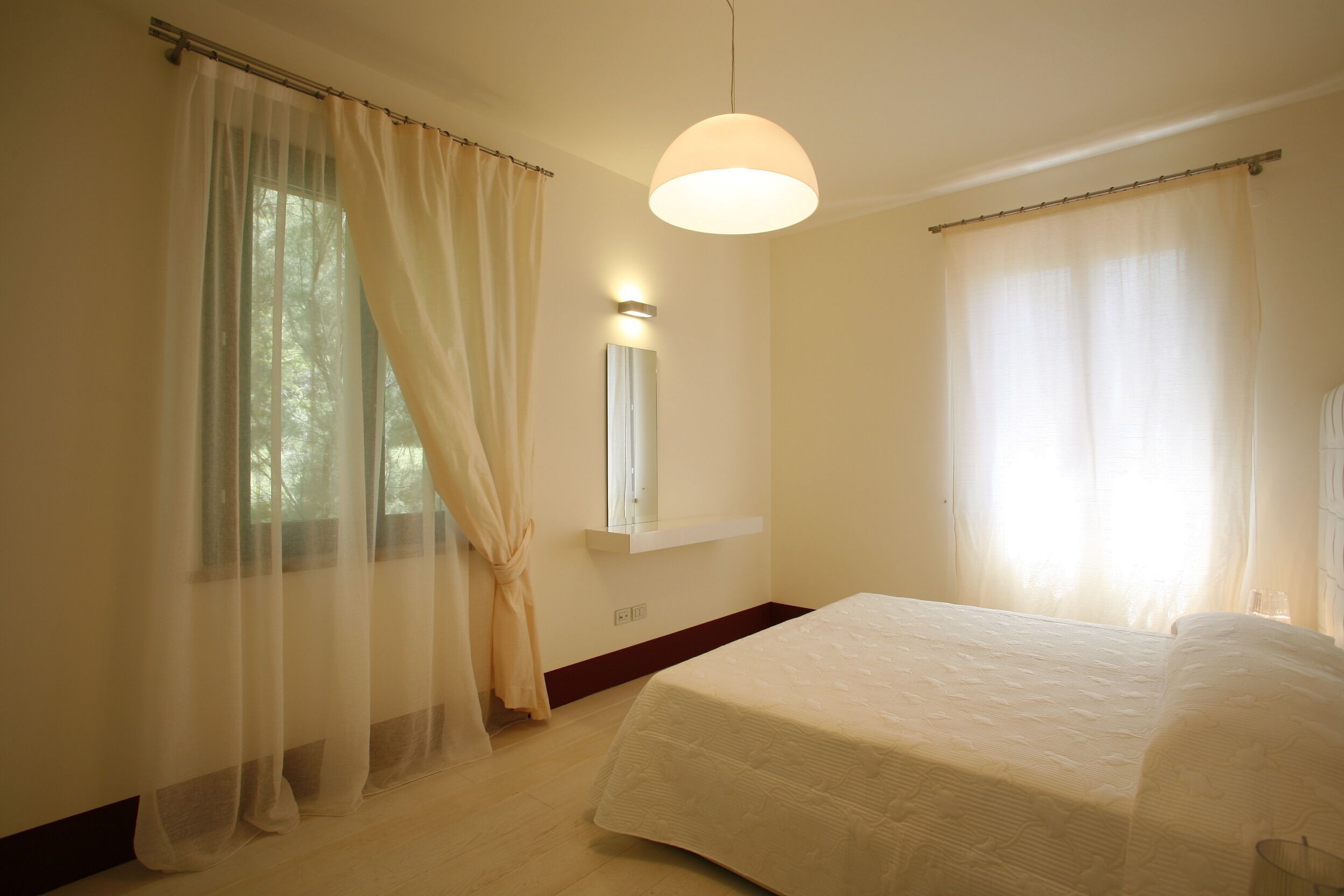 A bedroom with a bed, a window with sheer curtains, a wall-mounted mirror, and a pendant light.