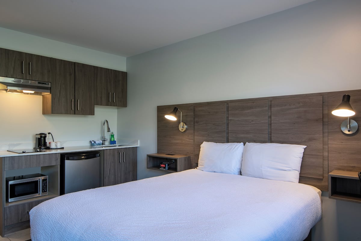 Une chambre d’hôtel moderne dotée d’un grand lit, d’une kitchenette équipée d’un four à micro-ondes, d’une machine à café et d’un lave-vaisselle, ainsi que d’une table de chevet avec une horloge.