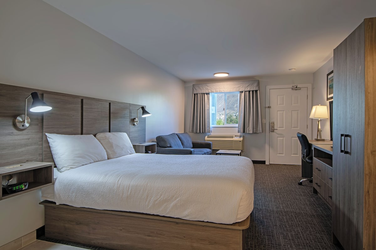 Une chambre d’hôtel avec un grand lit, un bureau, une chaise, une lampe de bureau, une table de chevet et une fenêtre avec des rideaux.